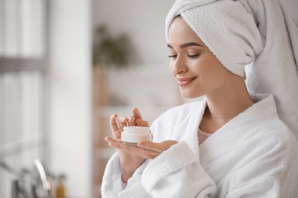Woman in a bathrobe and head towel scooping moisturizer from a jar after bathing, a gentle routine for dry skin