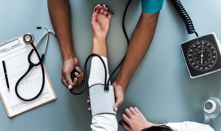 Clinician measuring a patient's blood pressure with a cuff and manual gauge beside a stethoscope and clipboard