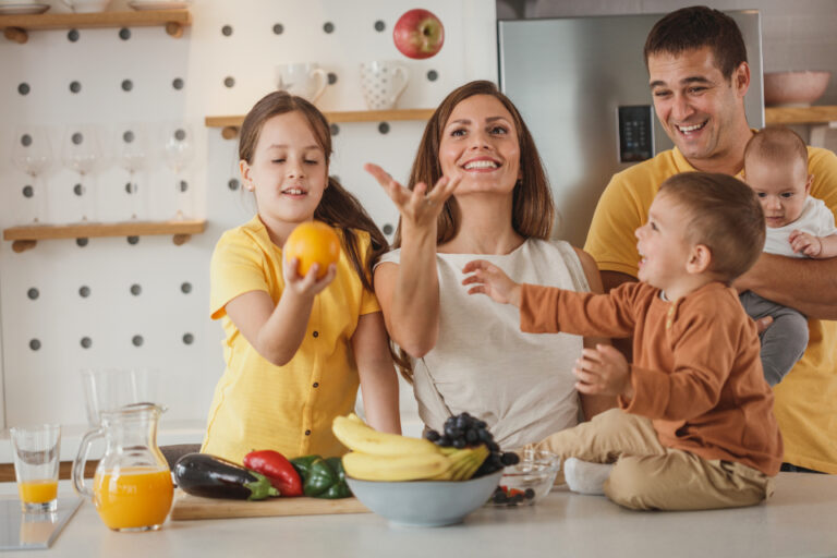 Smiling family of five around a kitchen counter with fruit, vegetables and orange juice, sharing a healthy meal