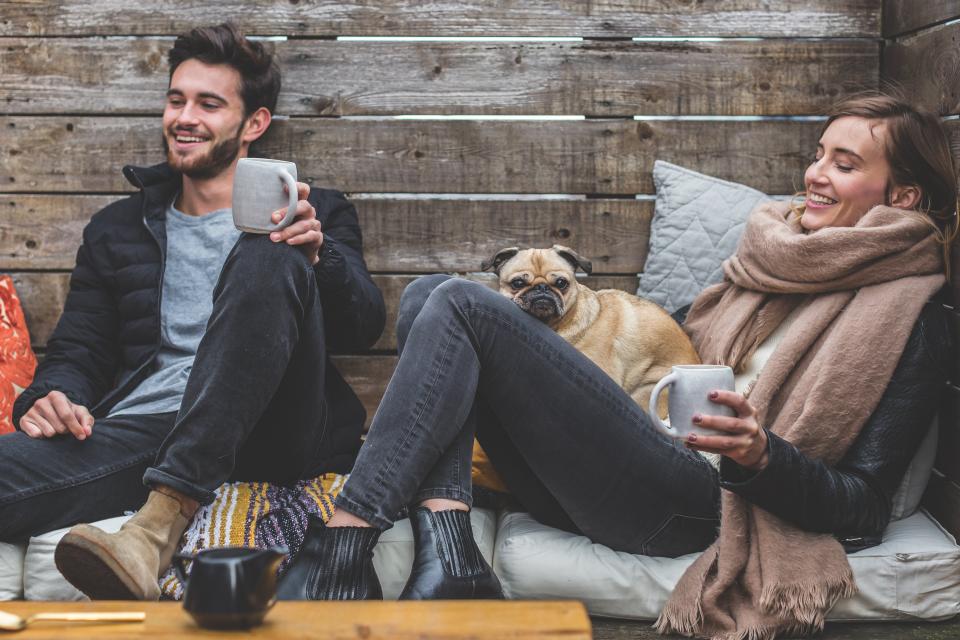 Relaxed couple laughing together with warm drinks and a pug, showing how rest and connection support intimacy