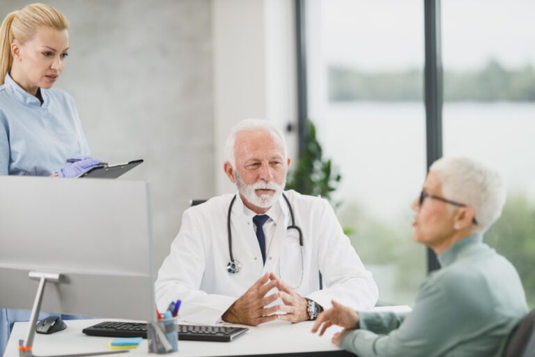 Doctor listening attentively to a patient across his desk in a bright office while a nurse reviews notes