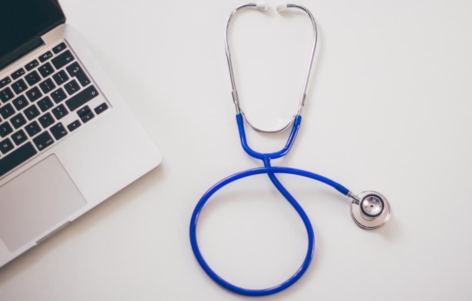 Stethoscope lying next to a laptop on a white desk, representing a telehealth visit with a doctor