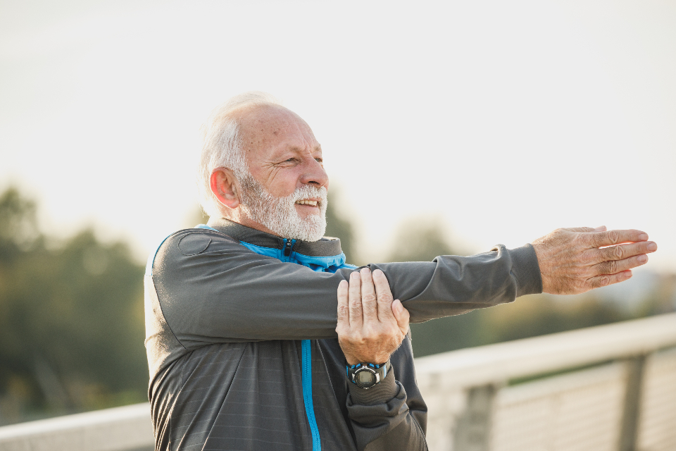 Smiling older man doing an arm stretch across his chest outdoors, practicing a simple home exercise