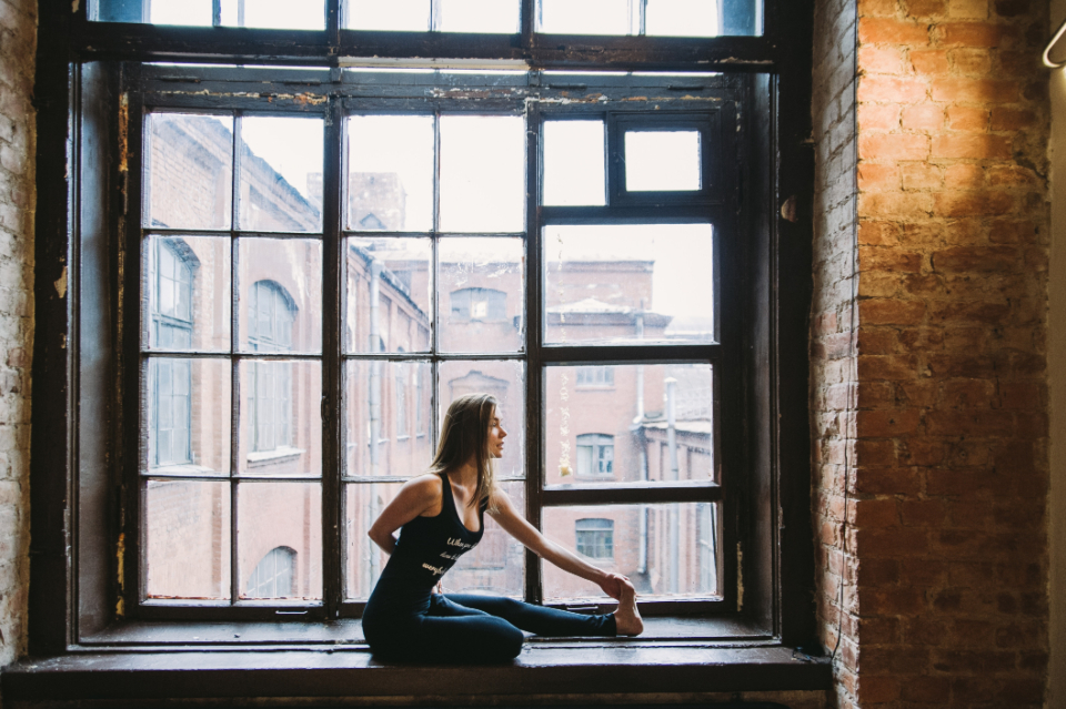 Woman gently stretching one leg while seated beside a large window, a form of gentle movement for chronic pain