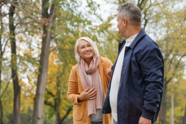 Smiling older woman in an orange coat chats with a man as they walk through an autumn park, staying active together