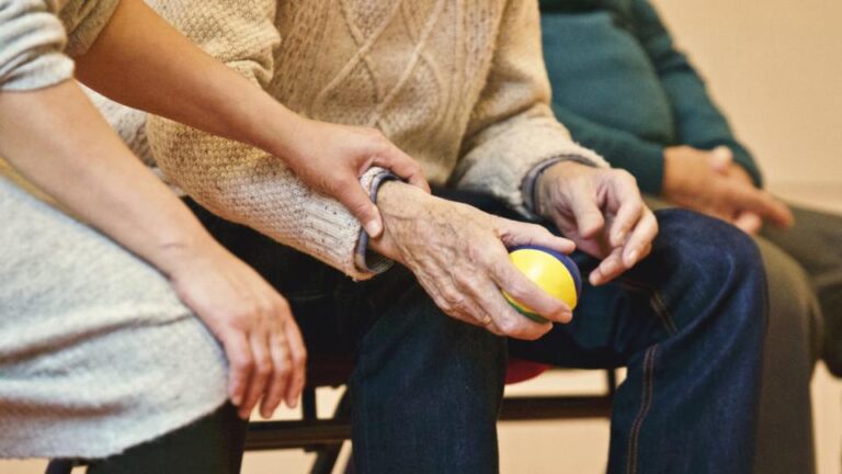 Older adult's hands resting on their knee, holding a small yellow ball, with a caregiver's hand on their arm