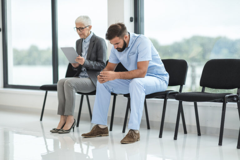 Two people waiting in a bright clinic waiting area, one holding a tablet and the other looking at a phone