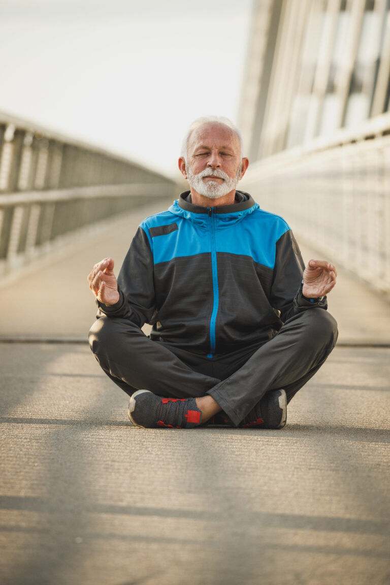 Older man sitting cross-legged outdoors with eyes closed and hands on his knees, practicing a calm breathing exercise