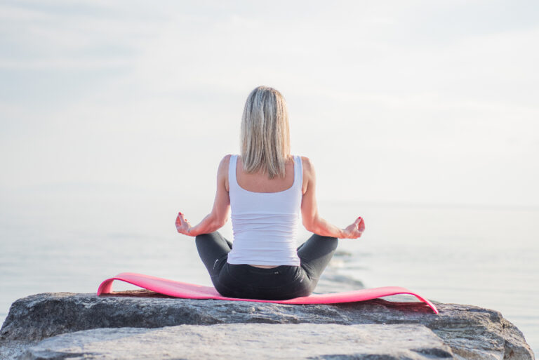 Woman sitting in a calm meditation pose on a rock by the sea, a quiet daily habit for mental wellbeing