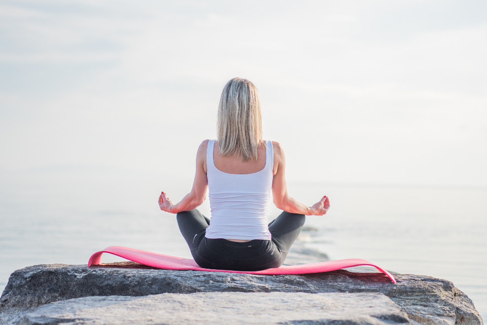 Woman sitting in a calm meditation pose on a rock by the sea, a quiet daily habit for mental wellbeing