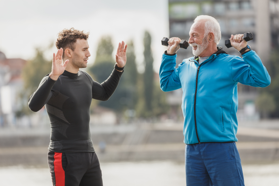 Older man lifting light dumbbells while a trainer guides him outdoors, building strength safely