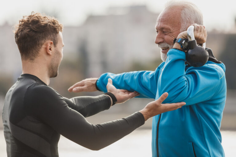 Fitness trainer guiding an older man through an arm and shoulder stretch outdoors during an exercise session
